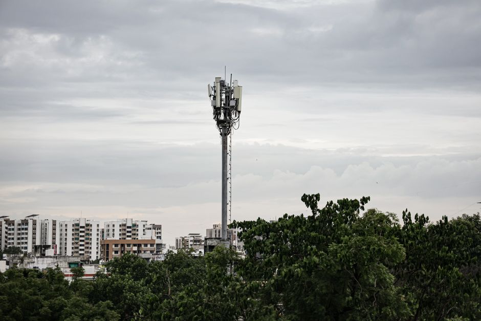 Cell tower rising above trees with urban high-rise buildings under a cloudy sky.